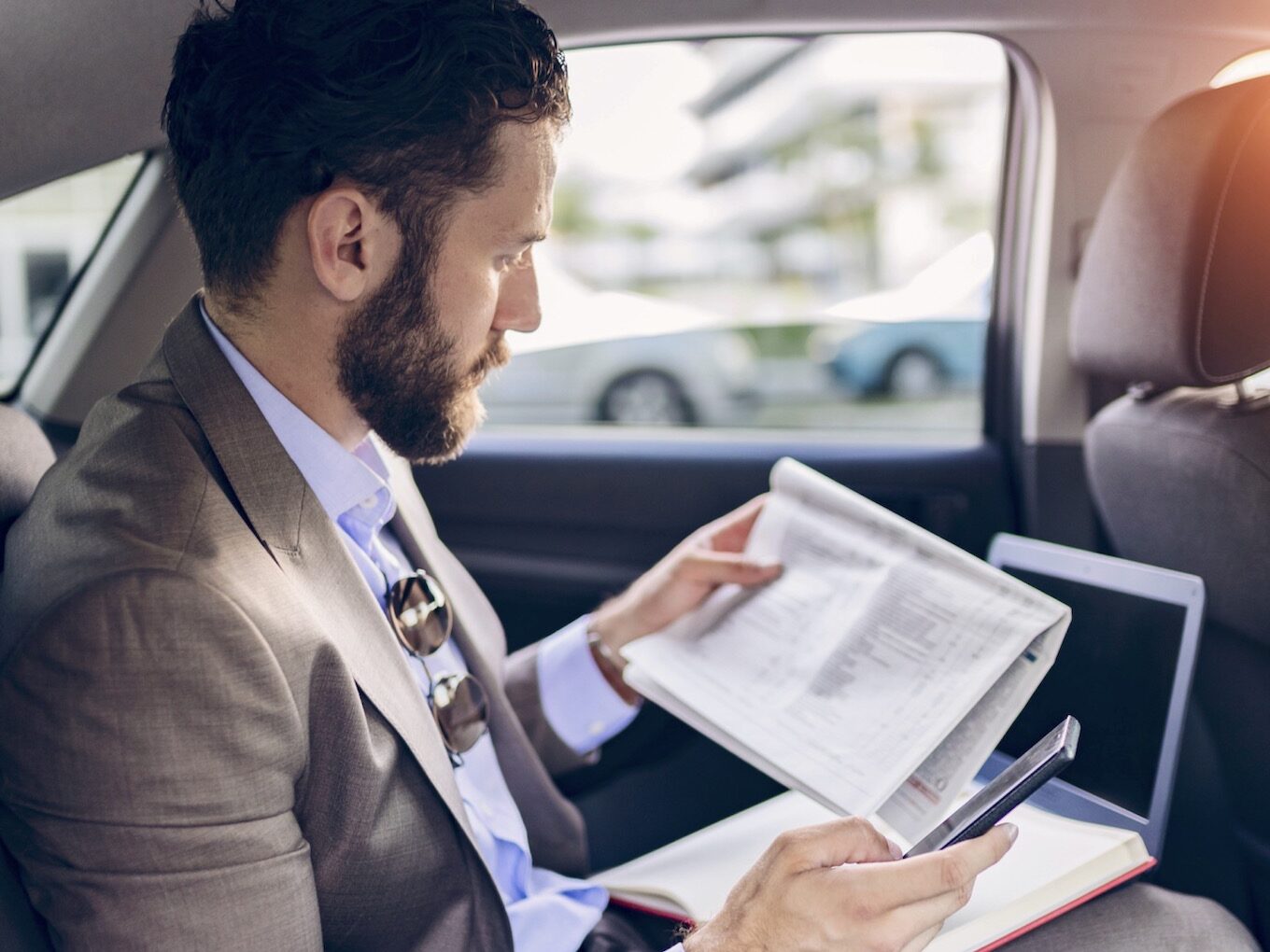 Confident man in full suit reading papers and using smart phone while sitting in the limo on the back seat with cityscape in the background. Transport, business trip, paperwork, people concept.