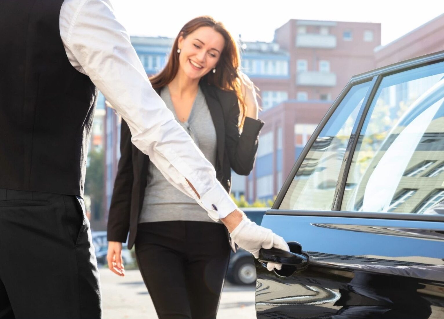 A chauffeur wearing white gloves opens a car door for a smiling woman who is approaching to enter the vehicle.