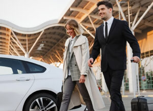A man and woman walk with luggage beside a white car outside a modern airport terminal.