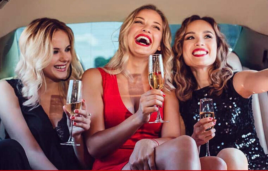 Featured_Three women dressed for a night out sit in a car, laughing, holding champagne glasses, and taking a selfie.