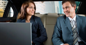 Two professionally dressed people sit in the backseat of a car, smiling at each other while one uses a laptop.