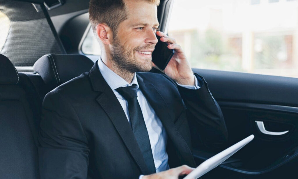 Featured_A man in a suit sits in the backseat of a car, smiling while talking on his phone and holding a tablet.
